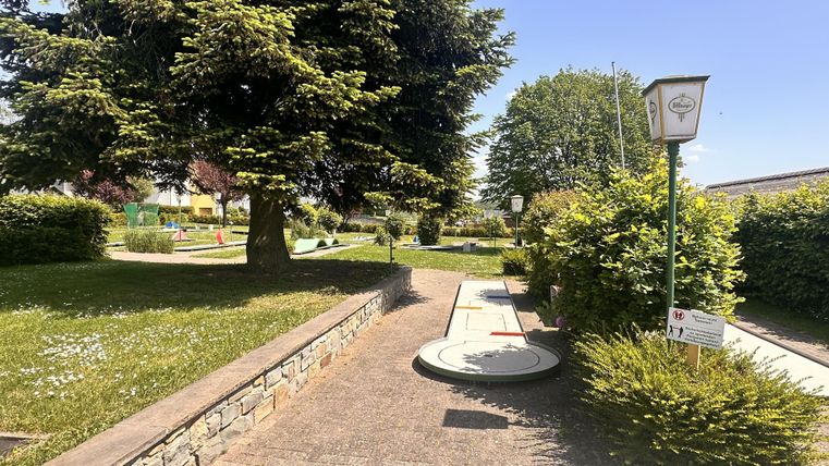 Ein gepflegter Minigolfplatz mit grünen Rasenflächen und Bäumen. Das Wetter ist sonnig und blau, ideal für einen Freizeitbesuch.