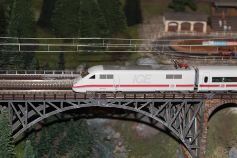 Un train modèle ICE passe sur un pont. À l'arrière-plan, on peut voir des arbres et un petit réseau ferroviaire.