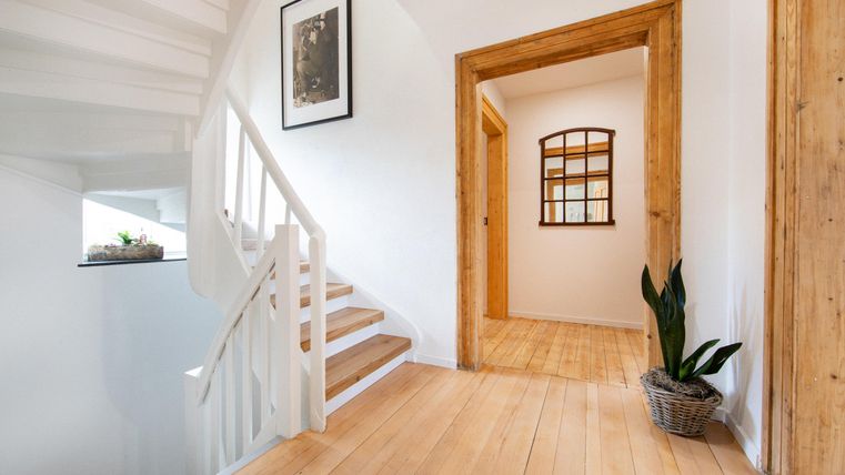 Ein heller Flur mit einer Treppe und Holzfußboden. Im Hintergrund sind offene Türen und ein Pflanzengefäß zu sehen.