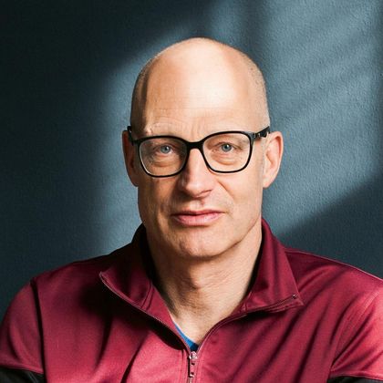 Ein Mann mit Brille und glattem Kopf in einem dunkelrot gefärbten Oberteil. Der Hintergrund ist in einem sanften Blau mit Lichtschatten.