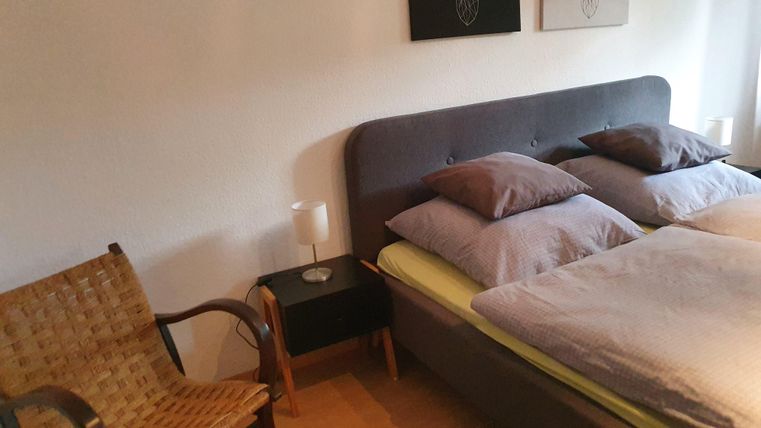 A cozy bedroom with a double bed and pillows. Next to the bed is a chair and a nightstand with a lamp.