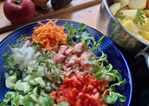Op een houten plank staat een bord met een salade met komkommers, paprika, uien, wortelen en vis; daarnaast staat een pot met aardappelen.