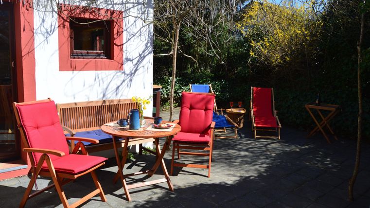 Ein gemütlicher Außenbereich mit roten Stühlen und einem Tisch. Im Hintergrund sind Pflanzen und ein helles Wetter zu sehen.