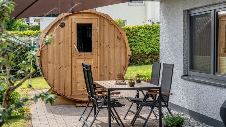 Ein kleines, rundes Holzhaus steht im Garten, umgeben von einer grünen Hecke. Vor dem Haus befinden sich ein Tisch und Stühle unter einem Sonnenschirm.