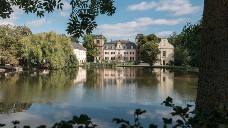 Eine malerische Landschaft mit einem ruhigen See und historischen Gebäuden im Hintergrund. Das Bild zeigt eine friedliche Atmosphäre mit schönem Wetter.