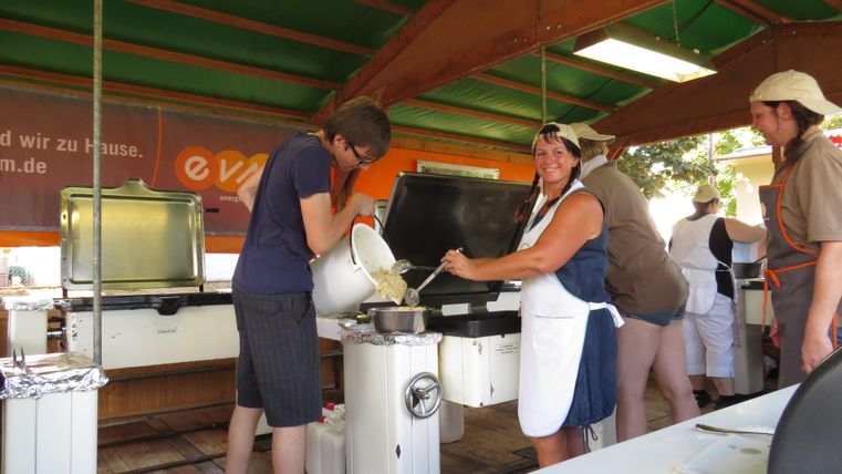 Un groupe de personnes cuisine dans une cuisine ouverte sous un toit en bois. Ils travaillent ensemble pour préparer un repas.