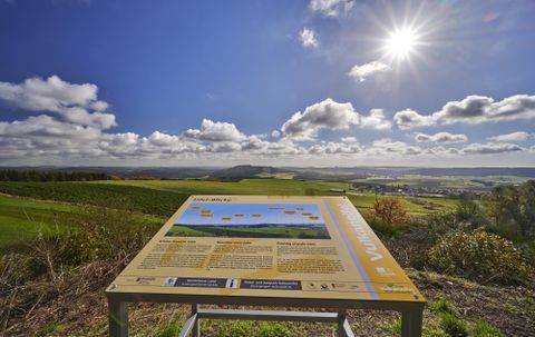 A viewing board on a hill with green fields and a clear sky. The sun is shining bright and there are a few clouds visible.