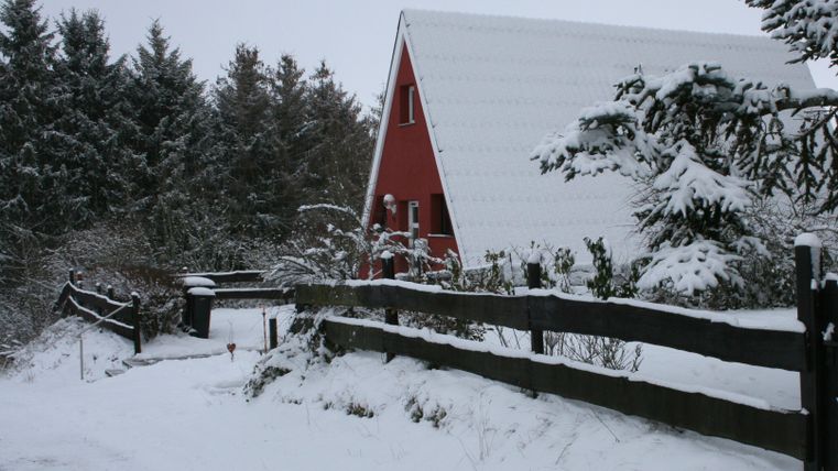 Une maison rouge dans un paysage enneigé. Le jardin est couvert de neige, et des arbres entourent le bâtiment.