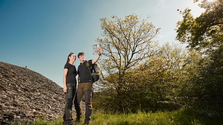 Zwei Personen machen ein Selfie vor einer großen Schieferhalde bei sonnigem Wetter; beide tragen Wanderkleidung, im Hintergrund sind Bäume mit frischem Laub zu sehen.