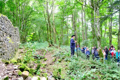 Een groep mensen luistert naar een spreker in het bos, terwijl groene bomen en planten de omgeving omringen. Op de achtergrond is een stenen muur zichtbaar.