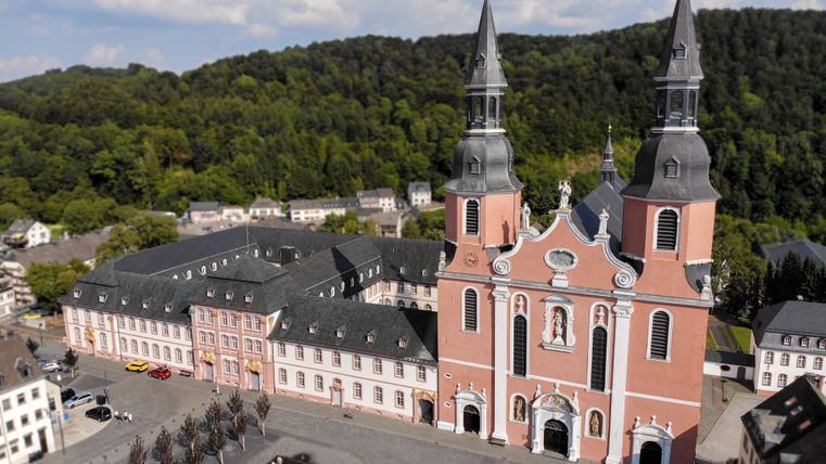 Eine beeindruckende Kirche mit zwei hohen Türmen steht im Vordergrund. Im Hintergrund sind grüne Hügel und ein Stadtplatz mit Bäumen zu sehen.