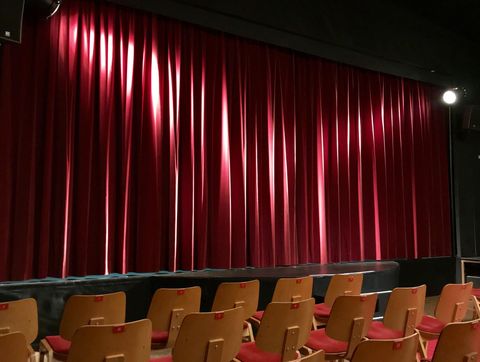Ein Theater mit roten Vorhängen und einer Reihe von Stühlen vor der Bühne. Die Atmosphäre ist gespannt und bereit für eine Vorstellung.