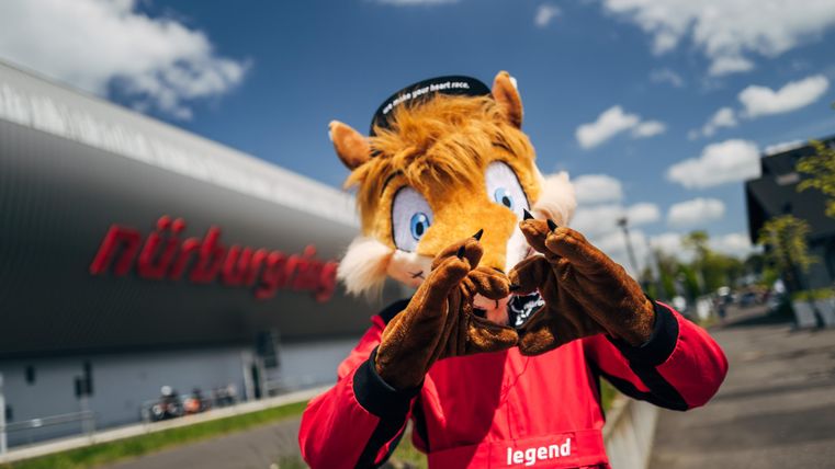 Ein Maskottchen in einem roten Overall macht ein Herzsymbol mit seinen Händen. Im Hintergrund ist das Gebäude des Nürburgrings zu sehen.