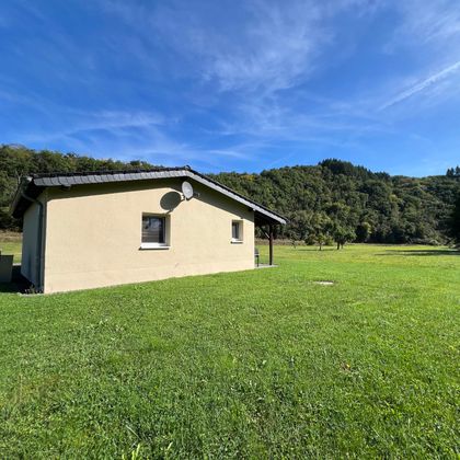 Une maison simple au milieu d'une prairie verte. À l'arrière-plan, on peut voir des collines douces et un ciel bleu clair.