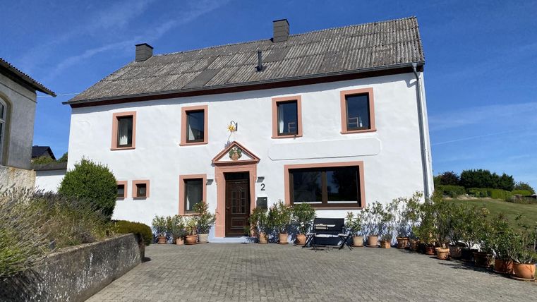 Ein hübsches Haus mit einer weißen Fassade und roten Fensterrahmen. Der Innenhof ist mit Blumentöpfen geschmückt und der Himmel ist klar und blau.