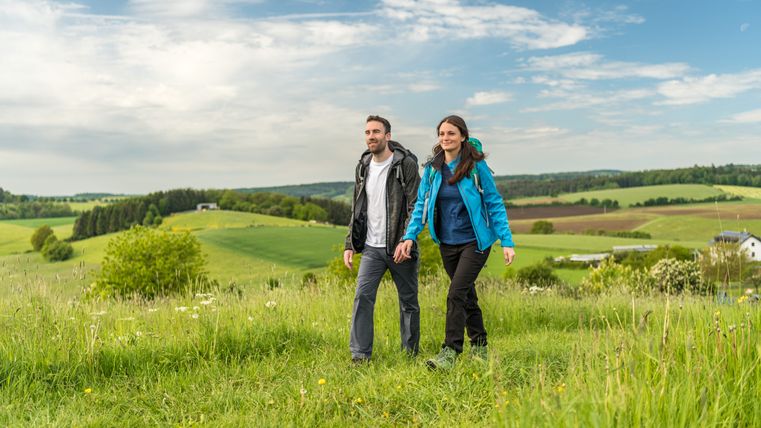A couple walks through a green landscape with hills and a blue sky.
