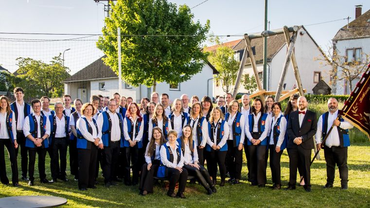 A large group of people is standing outside in a meadow. They are wearing blue vests and posing together with smiles.