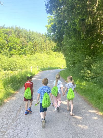 Vier Kinder mit grünen Rucksäcken gehen auf einem Waldweg spazieren.