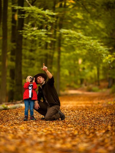 Un adulte montre quelque chose dans la forêt, tandis qu'un enfant le regarde attentivement. L'environnement est marqué par des couleurs automnales et des feuilles tombées.