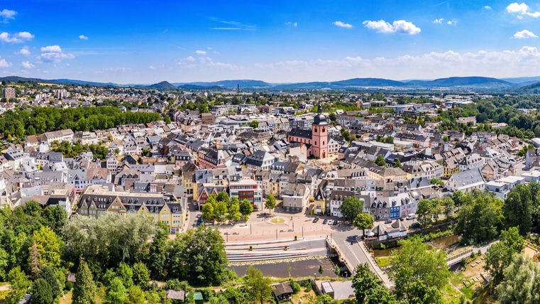 Eine Luftaufnahme einer Stadt mit vielen Häusern und einem großen Platz. Im Hintergrund sind Bäume und sanfte Hügel zu sehen, unter klarem, blauem Himmel.