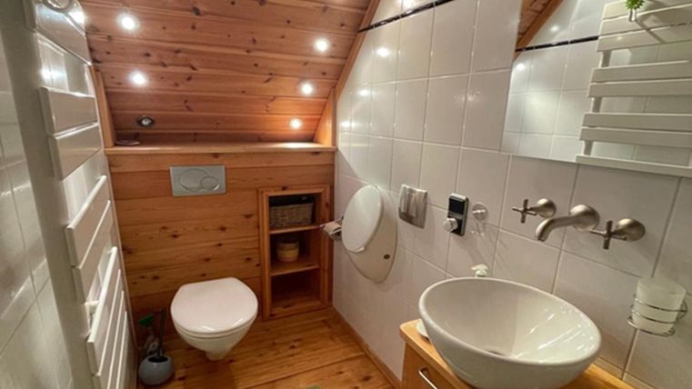 A modern bathroom with a wooden ceiling and light tiles. It features a toilet, a sink, and pleasant lighting.