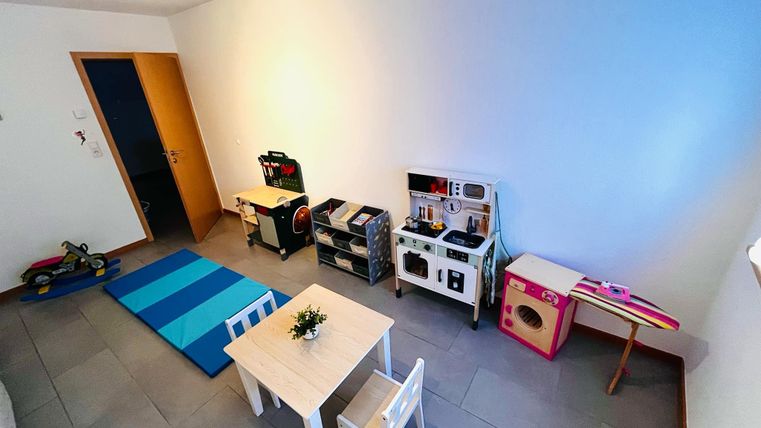 Een speelkamer met een kleine tafel en stoelen. Aan de muur staan keuken- en speelgereedschap en een blauwe tapijt ligt op de grond.