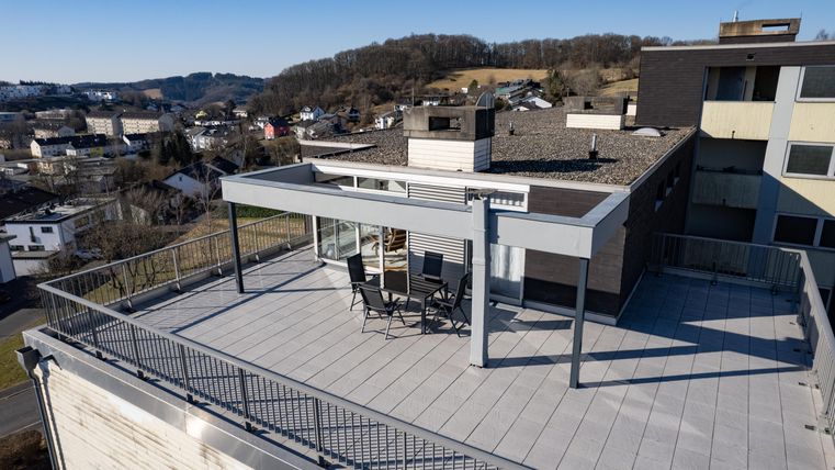 Een modern dakterras met uitzicht op een stadslandschap. Op het terras staan stoelen en een tafel onder een overkapping.