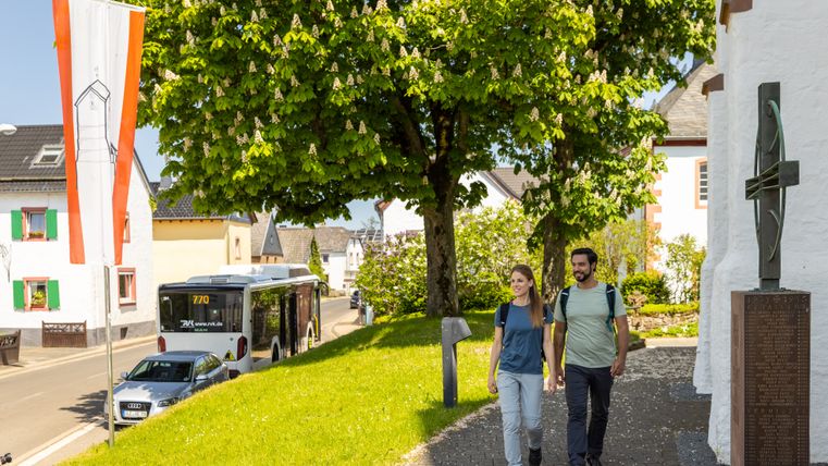 Deux randonneurs marchent sur un chemin pavé et passent devant une chapelle blanche ; à côté, un bus de randonnée « Eifelsteig » est garé au bord de la route. À l'arrière-plan, des arbres verdoyants, des maisons de village et un drapeau viennent agrémenter cette vue ensoleillée du village.