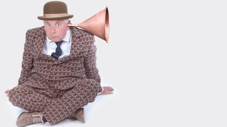 Ein Mann sitzt im Schneidersitz und trägt einen Anzug mit einem auffälligen Muster. An seinem Ohr ist ein großer Trichter angebracht, der an einen phonographischen Lautsprecher erinnert.