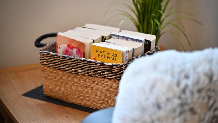 Ein Korb mit mehreren Büchern steht auf einem Tisch. Im Hintergrund ist eine Pflanze zu sehen.