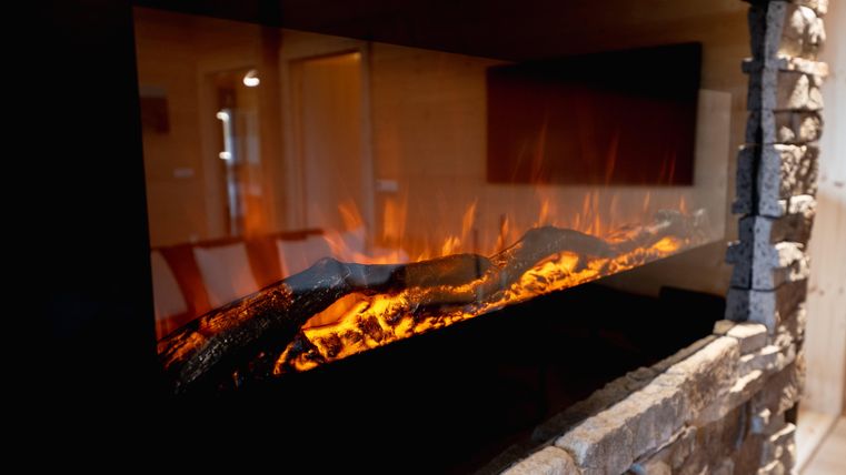 Ein moderner Kamin mit feurigen Flammen und hölzernen Scheiten. Die steinerne Umrandung verleiht dem Raum eine gemütliche Atmosphäre.
