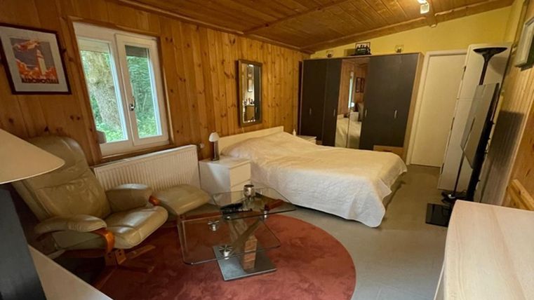 A cozy room with wooden walls and a bed. There is an armchair, a table, and large windows that let in light.