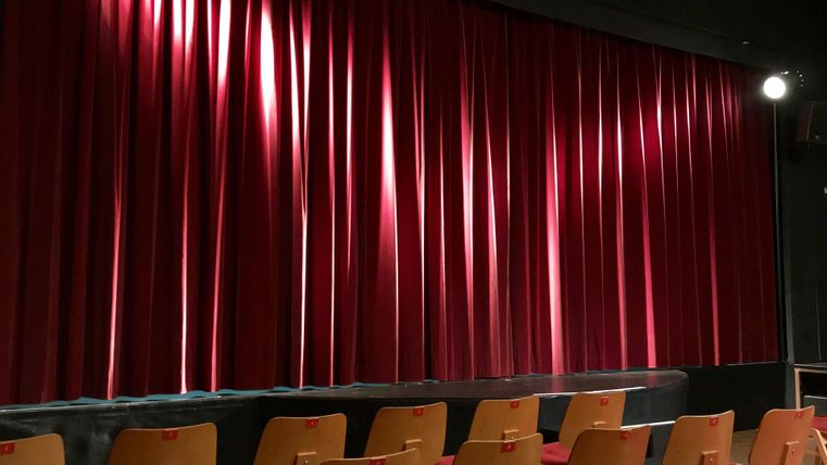 Ein Theater mit roten Vorhängen und einer leeren Sitzreihe. Das Licht beleuchtet die Vorhänge dezent.