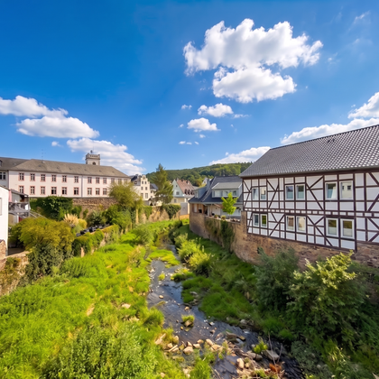 A picturesque landscape with a small stream, surrounded by green meadows. In the background, several charming houses stand beneath a bright blue sky with some clouds.