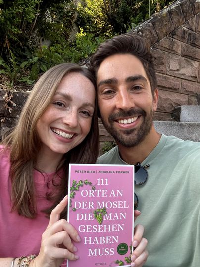 Ein lächelndes Paar sitzt im Freien und hält ein Buch mit dem Titel „111 Orte an der Mosel, die man gesehen haben muss“. Der Hintergrund zeigt grüne Pflanzen und eine steinerne Treppe.