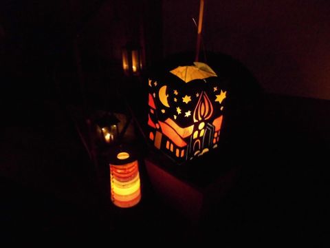Een kleurrijke lantaarn met feestelijke motieven straalt zachtjes in het donker. Naast staat een kleinere, gekleurde lantaarn die ook een warme gloed uitstraalt.