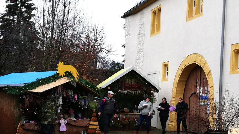 Ein einladender Markt mit zwei Ständen, umgeben von Bäumen, vor einem weißen Gebäude. Menschen schlendern vorbei und genießen die Atmosphäre.