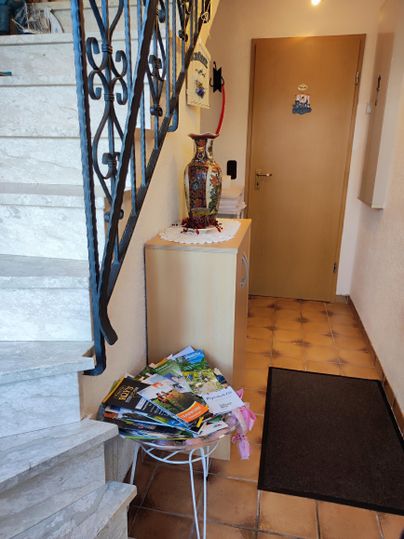 A hallway with a staircase, a side table, and a vase. On the table are numerous magazines and a doormat in front of the door.