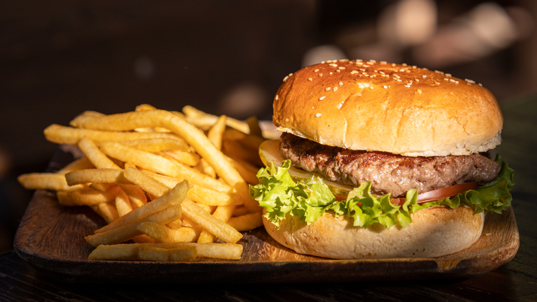 Ein saftiger Burger mit Sesambrötchen, Salat und Tomate liegt auf einem Holzbrett. Daneben befinden sich knusprige Pommes frites.