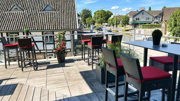 Eine moderne Terrasse mit Tischen und Stühlen in einem Restaurant. Im Hintergrund sind Häuser und Bäume zu sehen, unter einem klaren blauen Himmel.