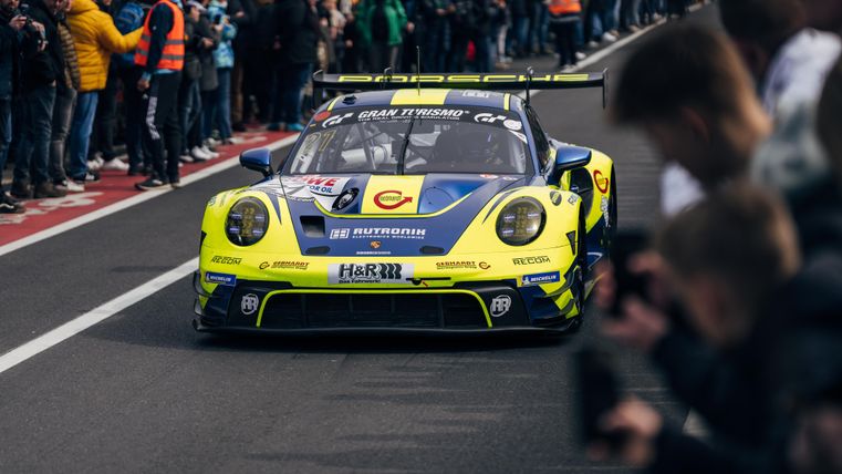 A race car is driving on the track, surrounded by enthusiastic spectators. The crowd is applauding and cheering.