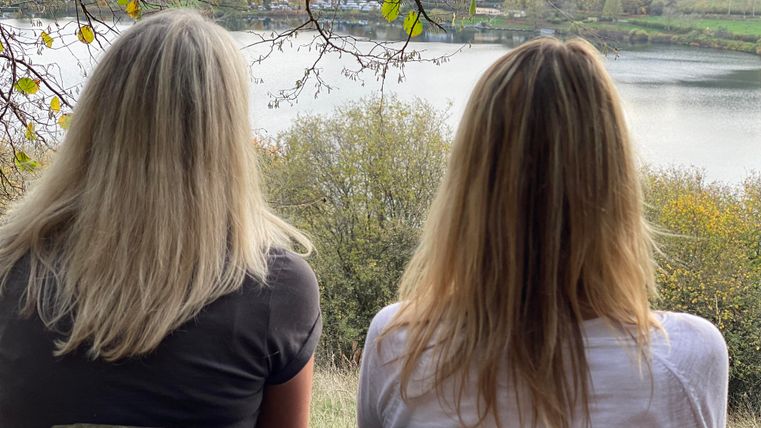 Twee vrouwen zitten op een bank en kijken naar een rustige vijver. Op de achtergrond zijn bomen en heuvels te zien.