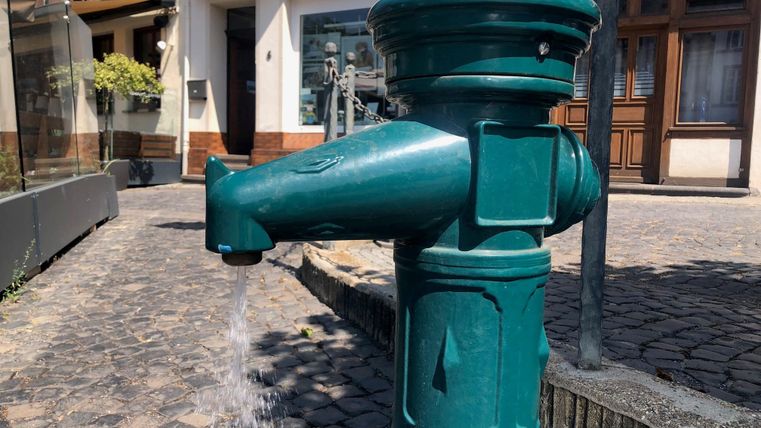 Un hydrant d'eau vert sur un pavé dans une ville. L'eau jaillit de l'hydrant et il y a quelques bâtiments commerciaux en arrière-plan.