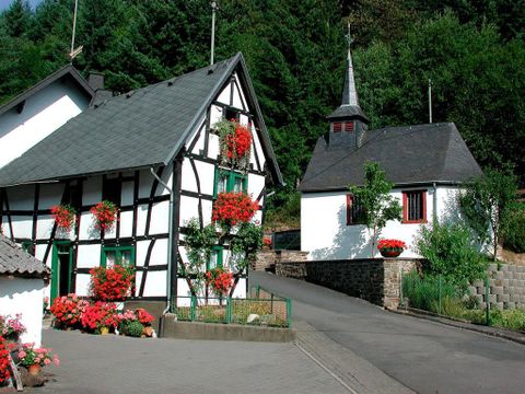 A picturesque village with traditional half-timbered houses and a small church. Colorful flowers adorn the buildings, and the surroundings are green and inviting.