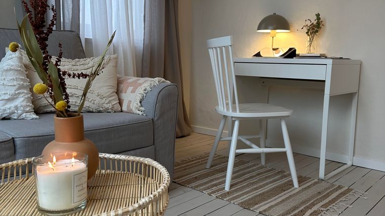 Een gezellige woonkamer met een grijze bank, een witte bureau en een stoel. Een tafel met een kaars en een plant maakt de ontspannen sfeer compleet.