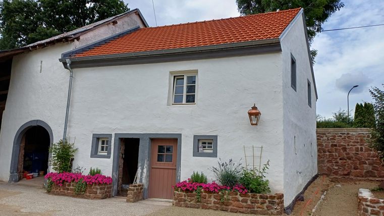A charming white house with a red roof and blooming plants in the front yard. The surroundings are calm and well-maintained.