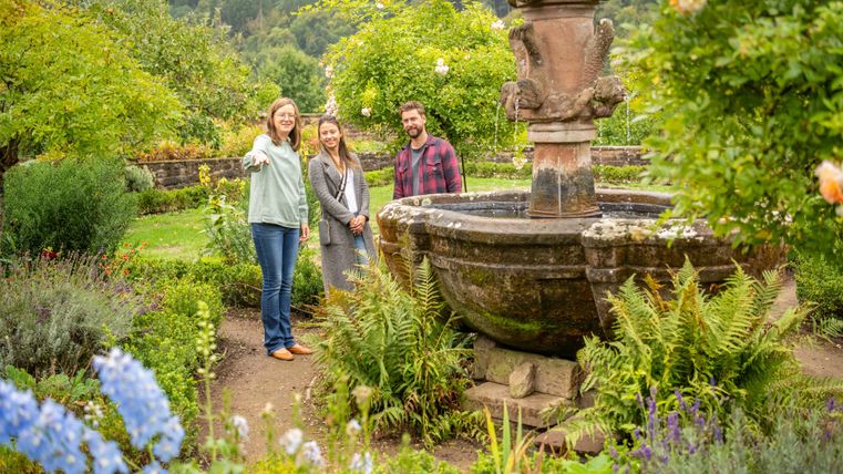 Drie personen in een mooie tuin staan om een historisch fontein. Rondom zijn groene planten en kleurrijke bloemen.