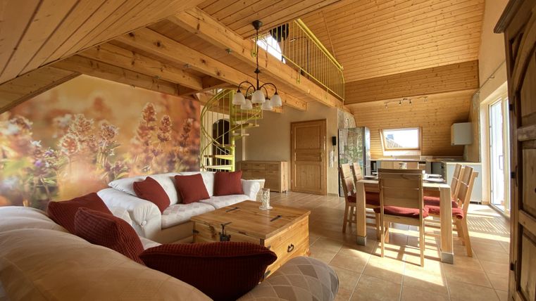 A cozy living room with a wooden ceiling and large windows. There is a sofa, a dining table, and a warm atmosphere.