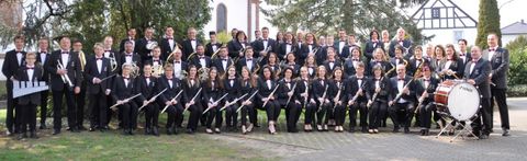 Un grand groupe de musiciens se tient à l'extérieur et joue différents instruments. Tous portent des costumes noirs élégants et sourient à la caméra.