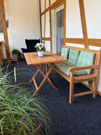 Un coin salon confortable sur un balcon avec une table en bois et un banc. Sur la table se trouve un petit vase à fleurs et en arrière-plan, des plantes sont visibles.